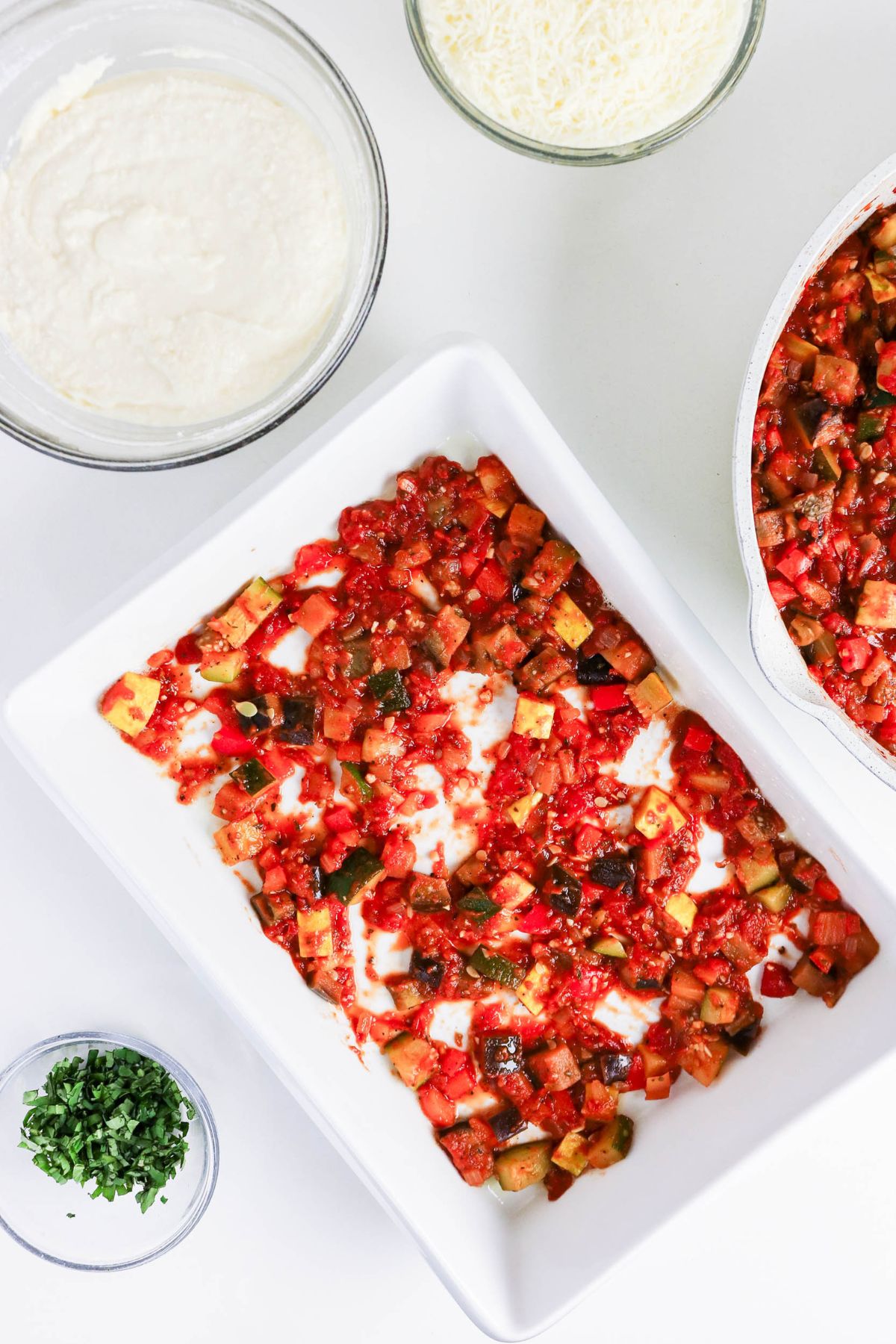 A baking dish with a layer of chopped tomato and vegetable mixture, surrounded by bowls of cheese, ricotta mixture, and chopped herbs on a white surface.