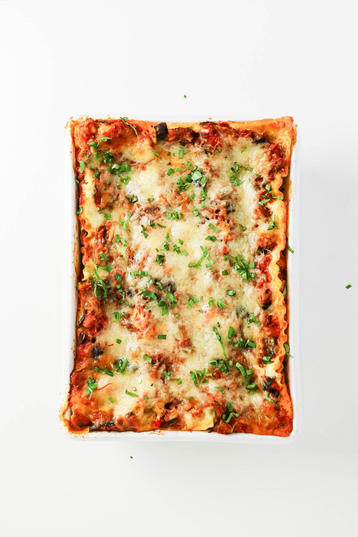 Rectangular baking dish filled with baked lasagna, topped with melted cheese and chopped herbs, photographed from above on a white background.