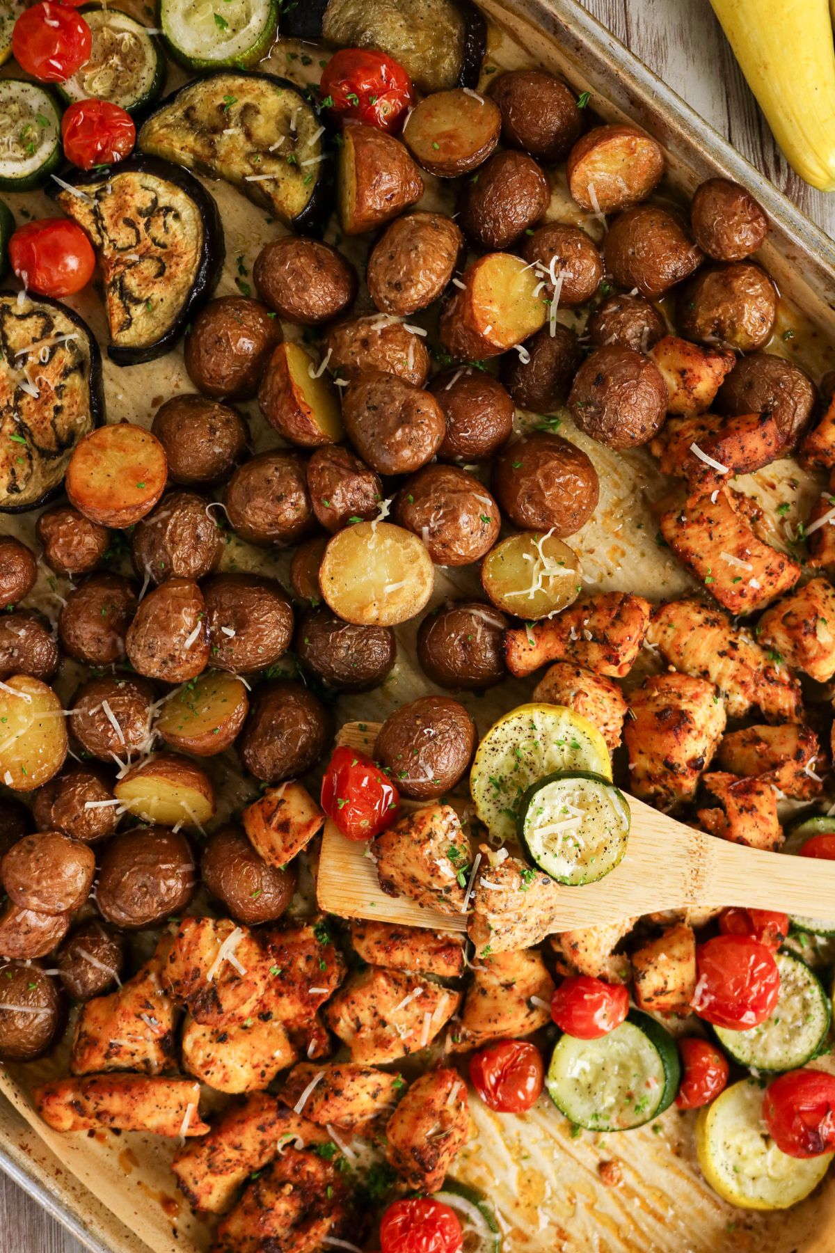 A sheet pan with roasted chicken, baby potatoes, zucchini, eggplant, and cherry tomatoes, garnished with herbs and grated cheese.
