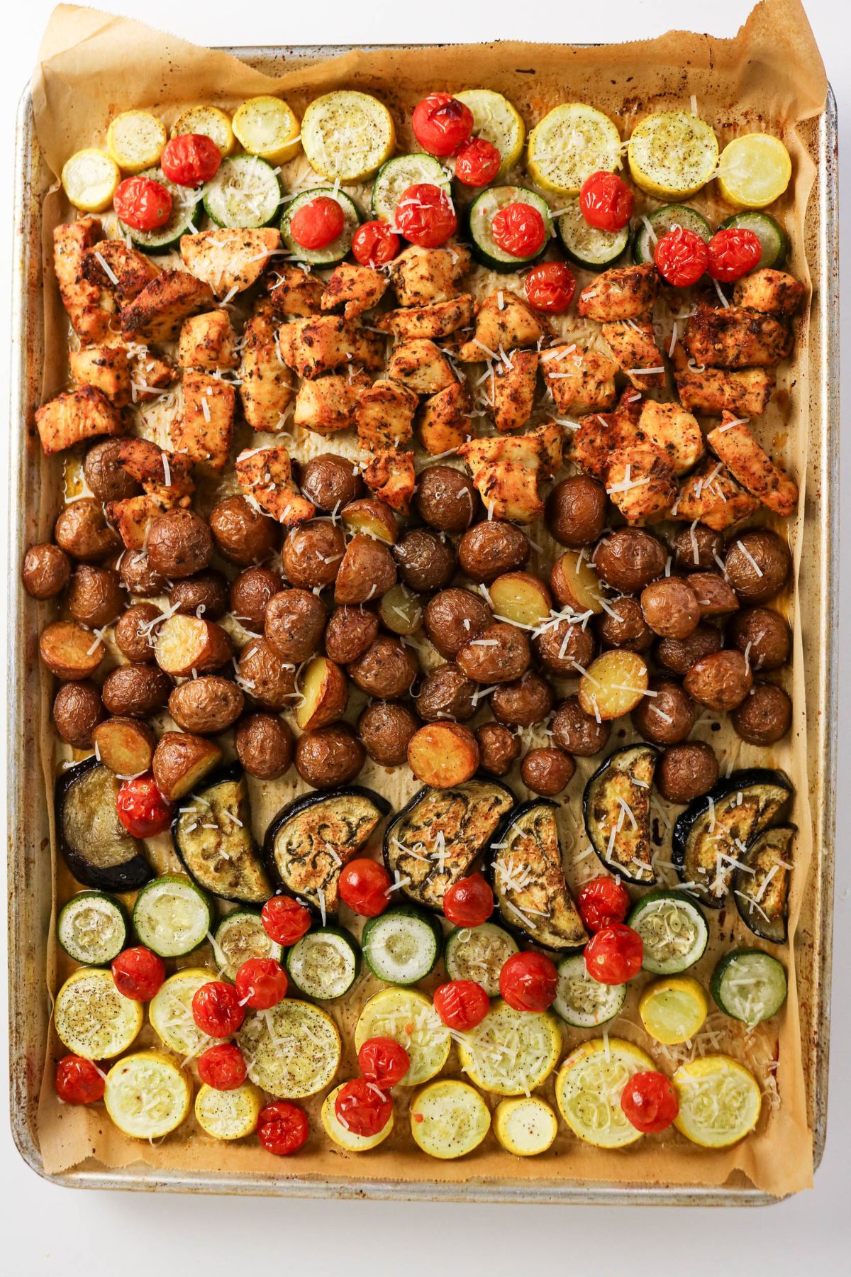 Sheet pan with baked chicken pieces, cherry tomatoes, zucchini, yellow squash, baby potatoes, and eggplant, all seasoned and sprinkled with grated cheese on parchment paper.