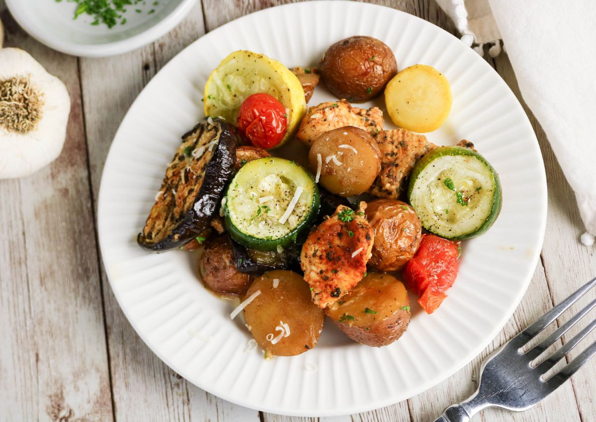 A white plate with roasted chicken, baby potatoes, zucchini, yellow squash, eggplant, cherry tomatoes, and grated cheese, on a wooden table with a fork and garlic nearby.