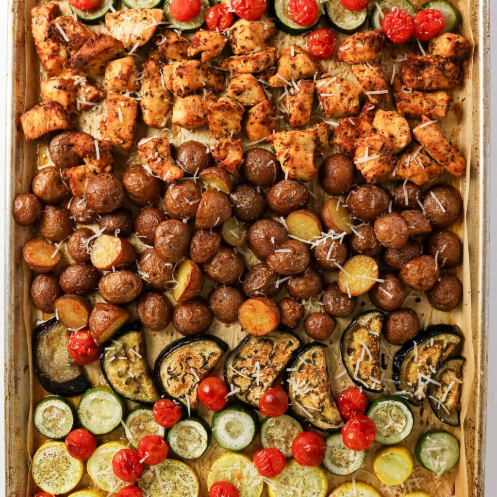 Sheet pan with rows of baked chicken pieces, baby potatoes, cherry tomatoes, zucchini, and eggplant, topped with shredded cheese.