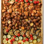 Sheet pan with rows of baked chicken pieces, baby potatoes, cherry tomatoes, zucchini, and eggplant, topped with shredded cheese.