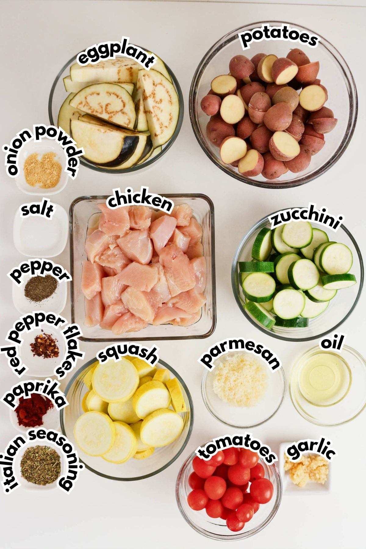 Bowls containing sliced eggplant, potatoes, chicken, zucchini, squash, grated parmesan, cherry tomatoes, olive oil, garlic, and small bowls of various spices are arranged on a white surface.