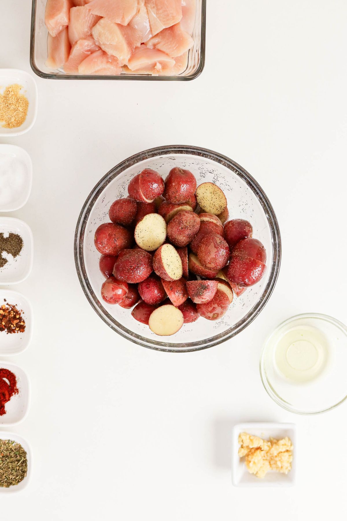 Overhead view of a bowl of halved red potatoes with seasoning, surrounded by diced raw chicken, oil, minced garlic, and small containers of seasonings on a white surface.