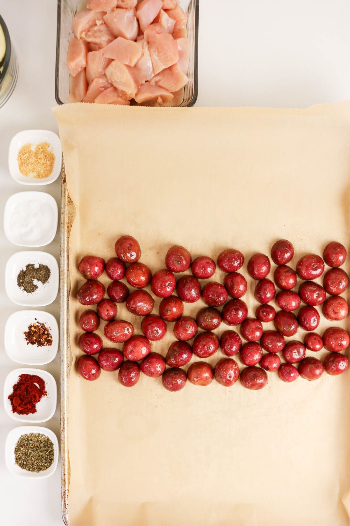 A baking sheet with parchment paper holds red potatoes. Surrounding the sheet are raw chicken pieces and small bowls of various spices and seasonings.