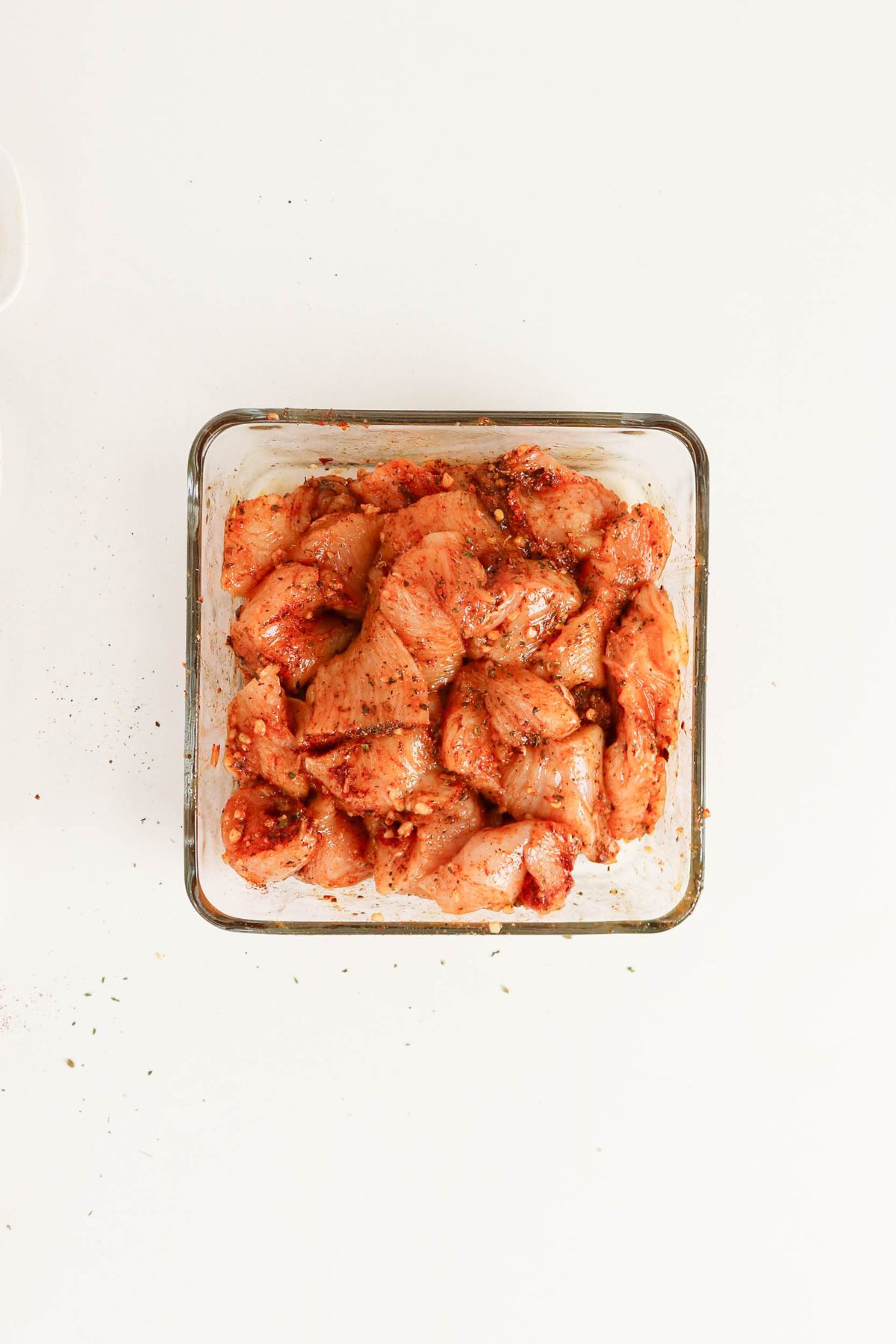 Glass container filled with raw, seasoned chicken pieces on a white surface; some spices are scattered around the container.
