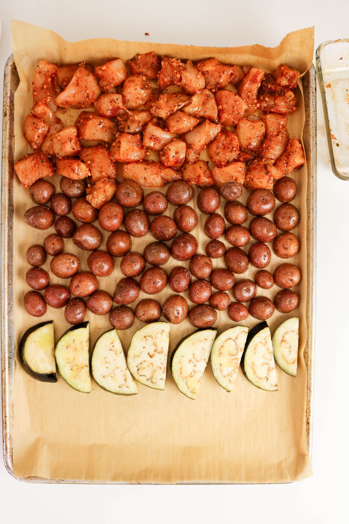 Sheet pan with rows of seasoned chicken pieces, whole baby potatoes, and sliced eggplant on parchment paper, ready to be cooked.