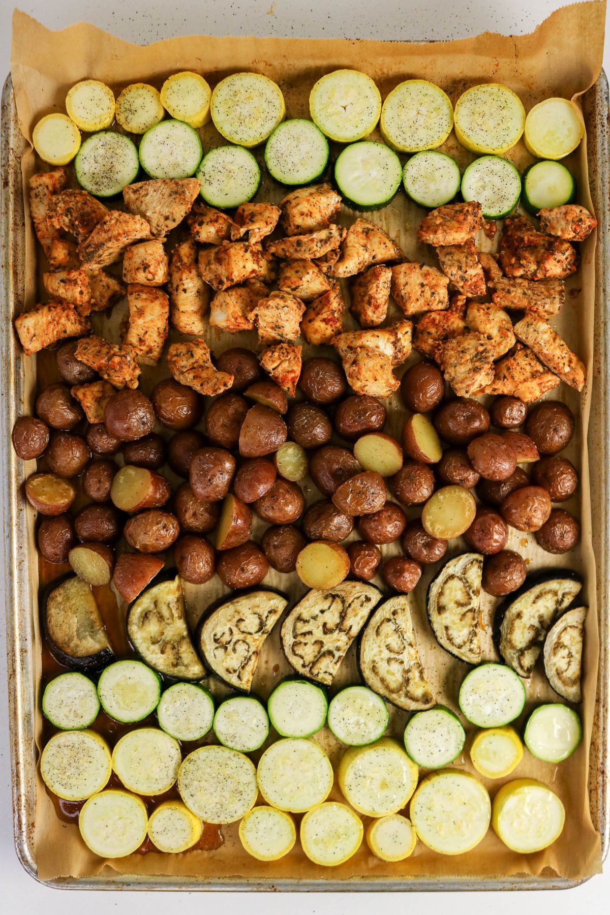 Baking sheet with rows of sliced zucchini, seasoned cooked chicken pieces, halved baby potatoes, and sliced eggplant on parchment paper.
