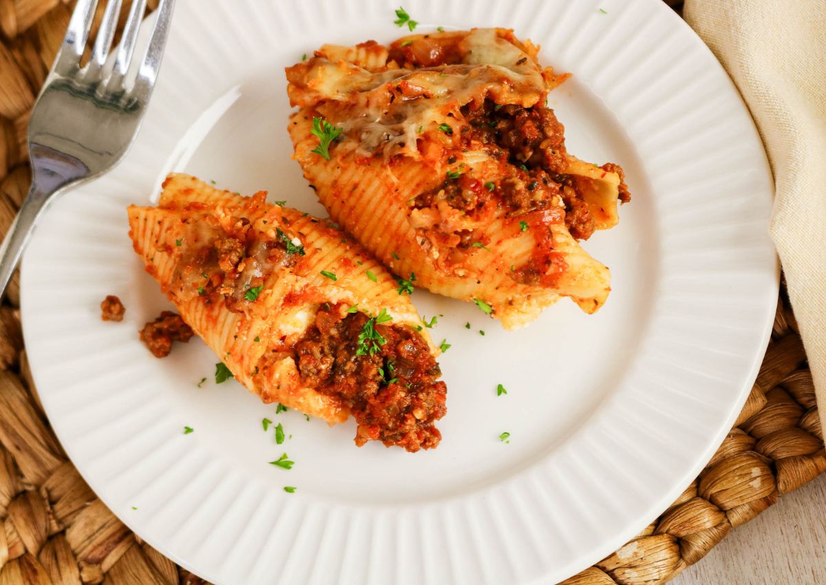 Three stuffed pasta shells with meat sauce and melted cheese on a white plate, garnished with chopped parsley, with a fork nearby.