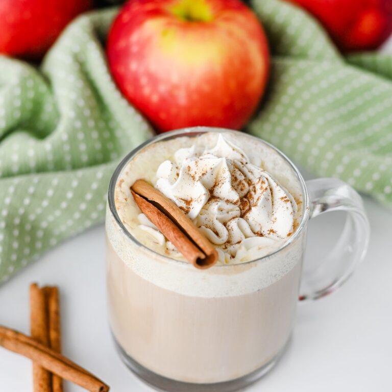 Apple chai latte in a glass mug with whipped cream, cinnamon stick, and apples in the background
