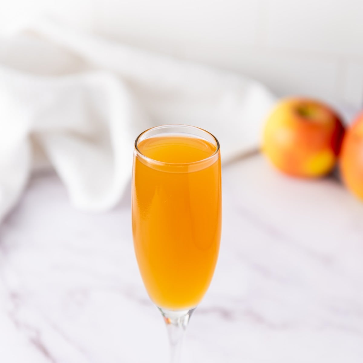 A champagne flute filled with orange-colored liquid sits on a marble surface, with apples and a white cloth blurred in the background.