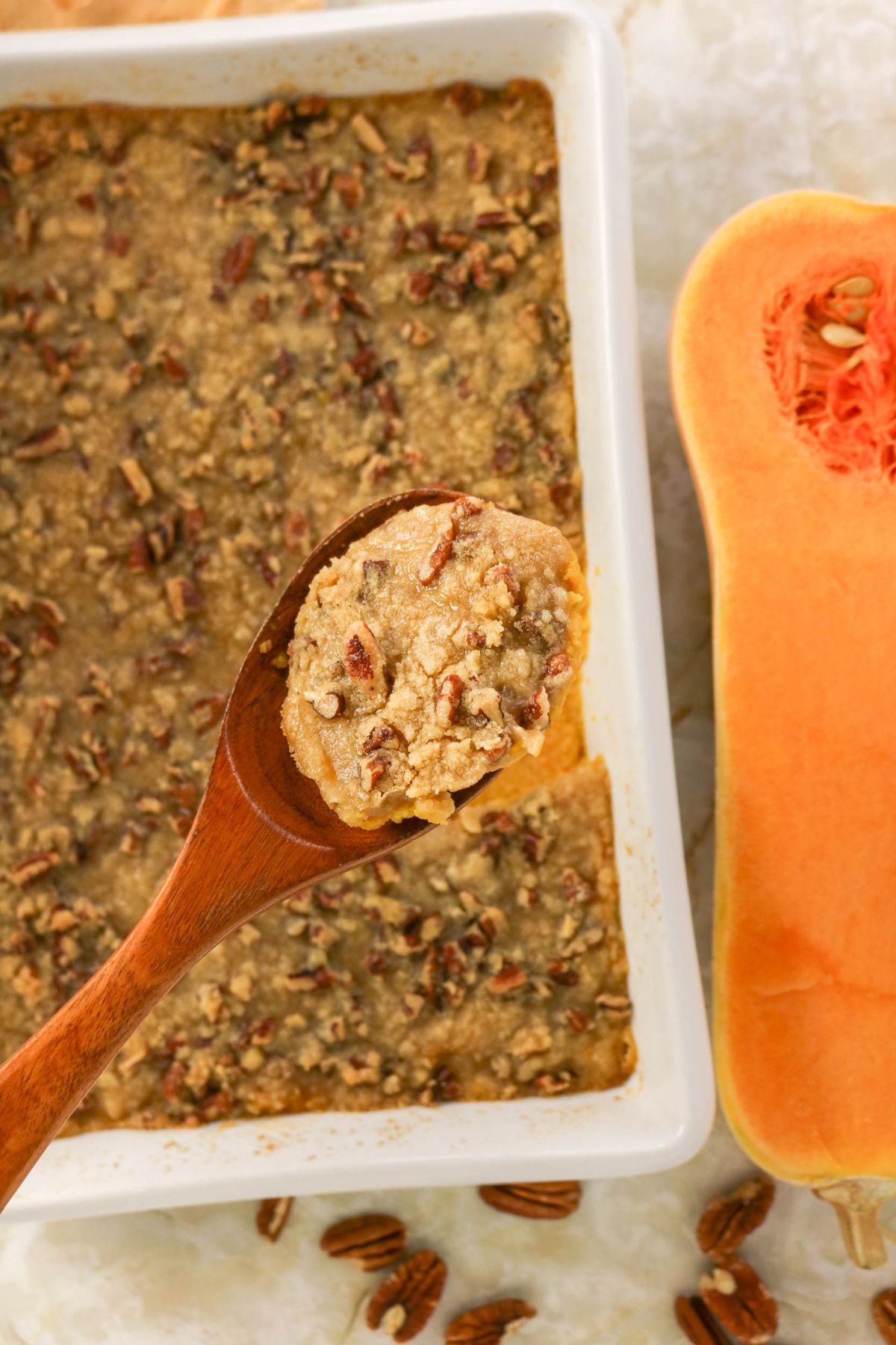 A wooden spoon lifts a serving of baked casserole topped with chopped pecans from a white dish, next to a halved butternut squash and scattered pecans.