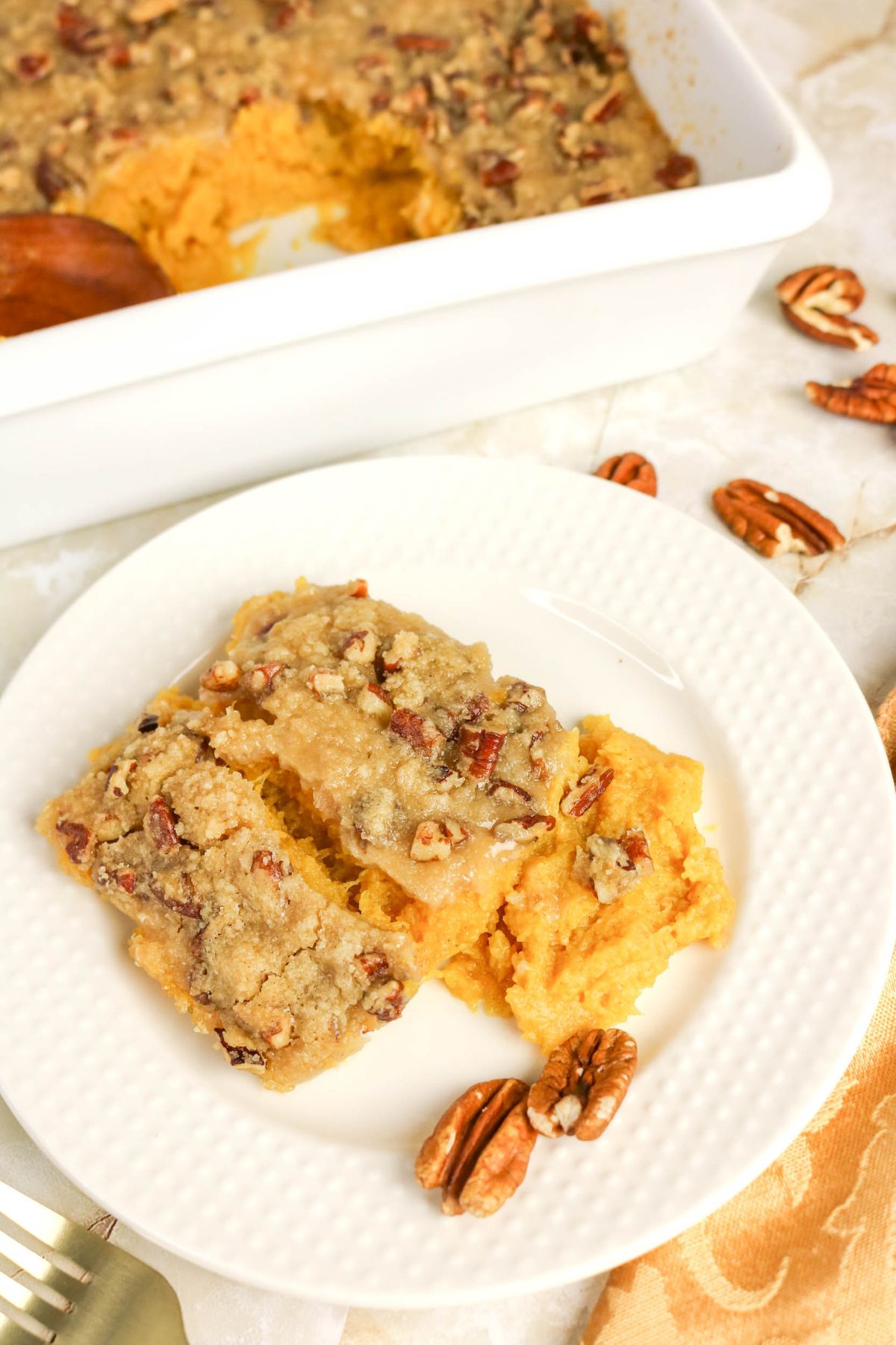 Two slices of sweet potato casserole with pecan crumble topping on a white plate, with whole pecans scattered nearby and the casserole dish in the background.
