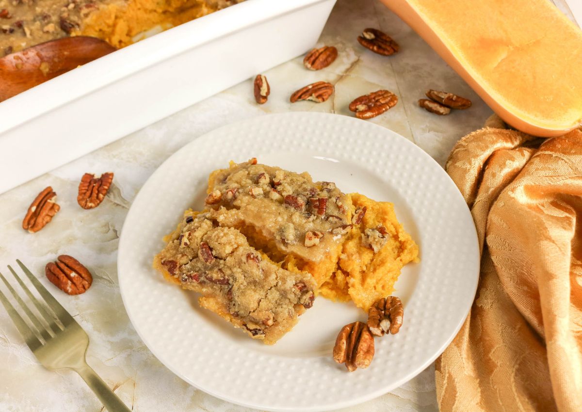 A white plate with two slices of sweet potato casserole topped with pecans, set near a casserole dish, pecans, fork, and orange napkin.