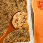 A wooden spoon holds a serving of pecan-topped casserole above a baking dish, with a halved butternut squash visible beside it.