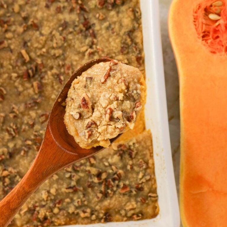 A wooden spoon holds a serving of pecan-topped casserole above a baking dish, with a halved butternut squash visible beside it.
