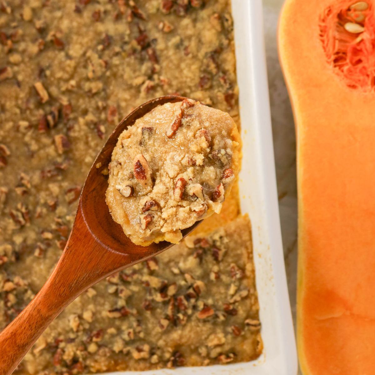 A wooden spoon holds a serving of pecan-topped casserole above a baking dish, with a halved butternut squash visible beside it.