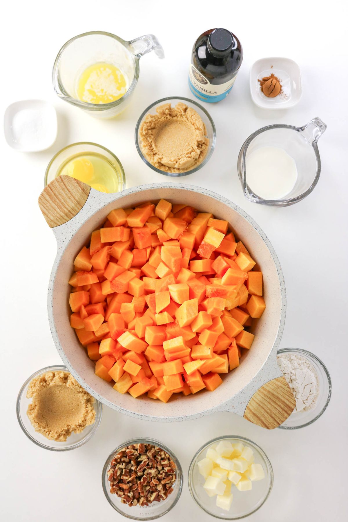 Chopped sweet potatoes in a bowl surrounded by brown sugar, pecans, melted butter, milk, vanilla, ground cinnamon, flour, and cubed butter, all in clear bowls on a white surface.