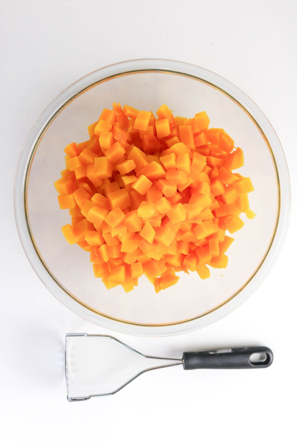 A glass bowl filled with diced orange squash sits on a white surface next to a metal potato masher with a black handle.