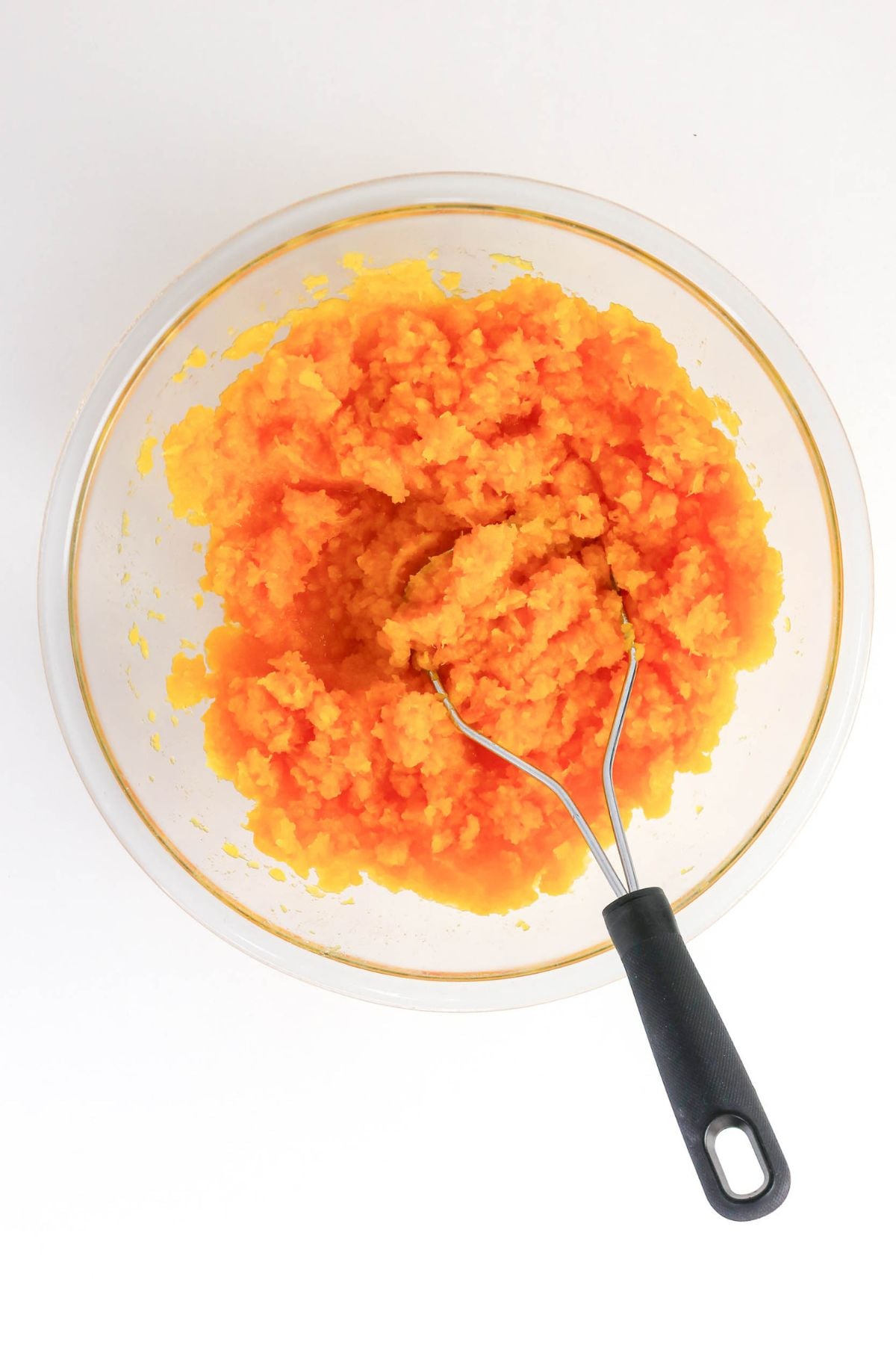 A glass bowl filled with mashed orange pumpkin, with a metal potato masher resting inside, set on a white background.