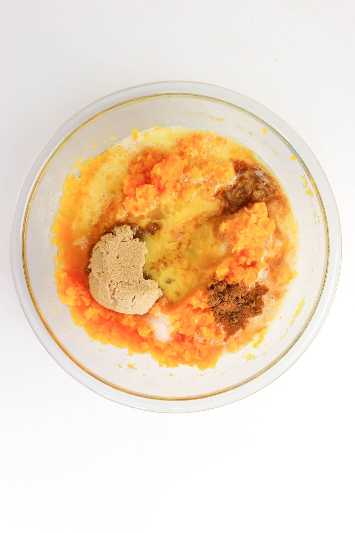 A clear bowl with mashed orange sweet potatoes, brown sugar, melted butter, milk, cinnamon, and other ingredients ready to be mixed.