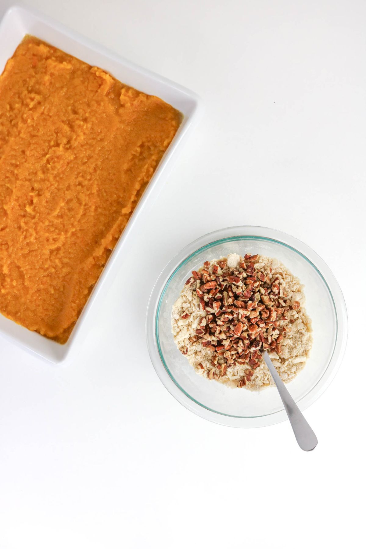 A rectangular dish of mashed sweet potatoes next to a bowl containing oats and chopped pecans with a spoon.