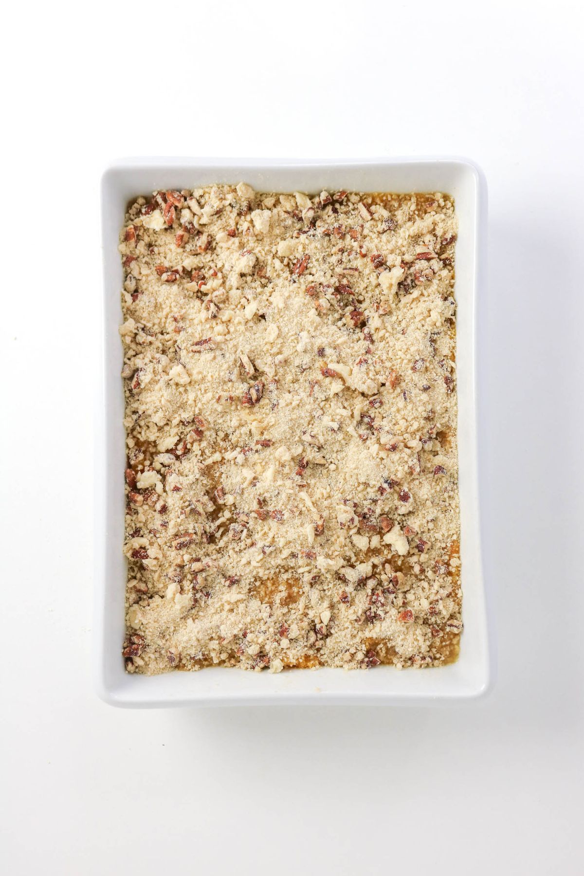 A white rectangular baking dish filled with an uncooked layered mixture topped with a crumbly, nutty streusel.