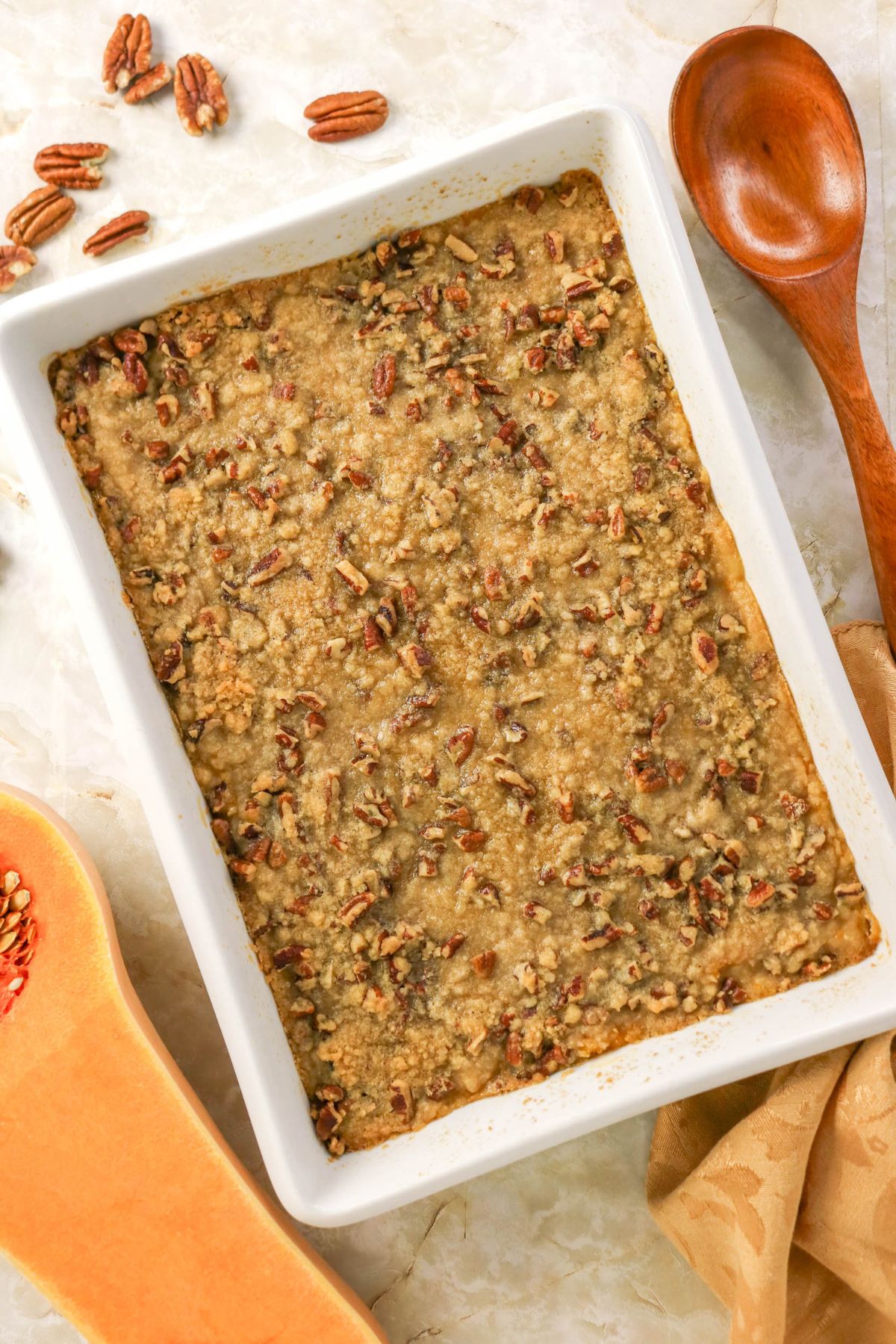 A baked casserole topped with chopped pecans in a white rectangular dish, placed on a light surface with a wooden spoon and scattered pecans nearby.