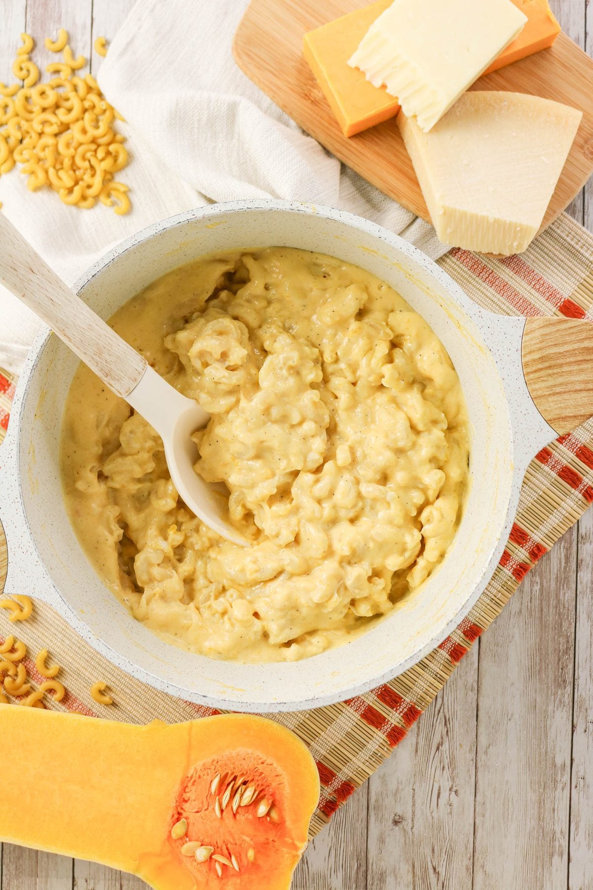 A pot of cooked macaroni and cheese with a white spoon, next to uncooked pasta, cheese blocks, and a sliced butternut squash on a wooden surface.