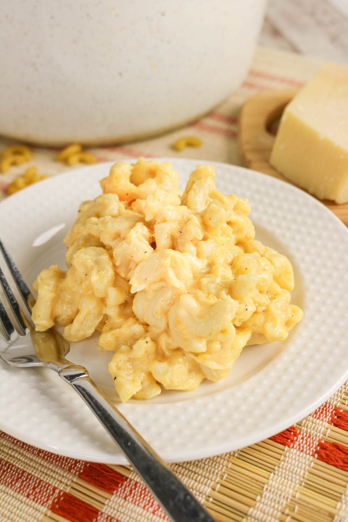 A serving of creamy macaroni and cheese on a white plate with a fork, set on a placemat near a block of cheese.