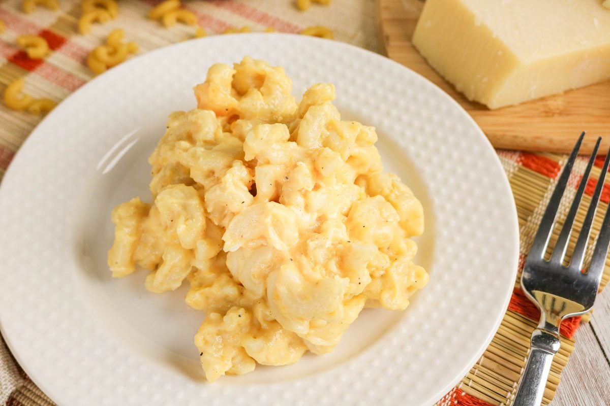 A serving of macaroni and cheese is on a white plate, next to a fork and a block of cheese on a cutting board.
