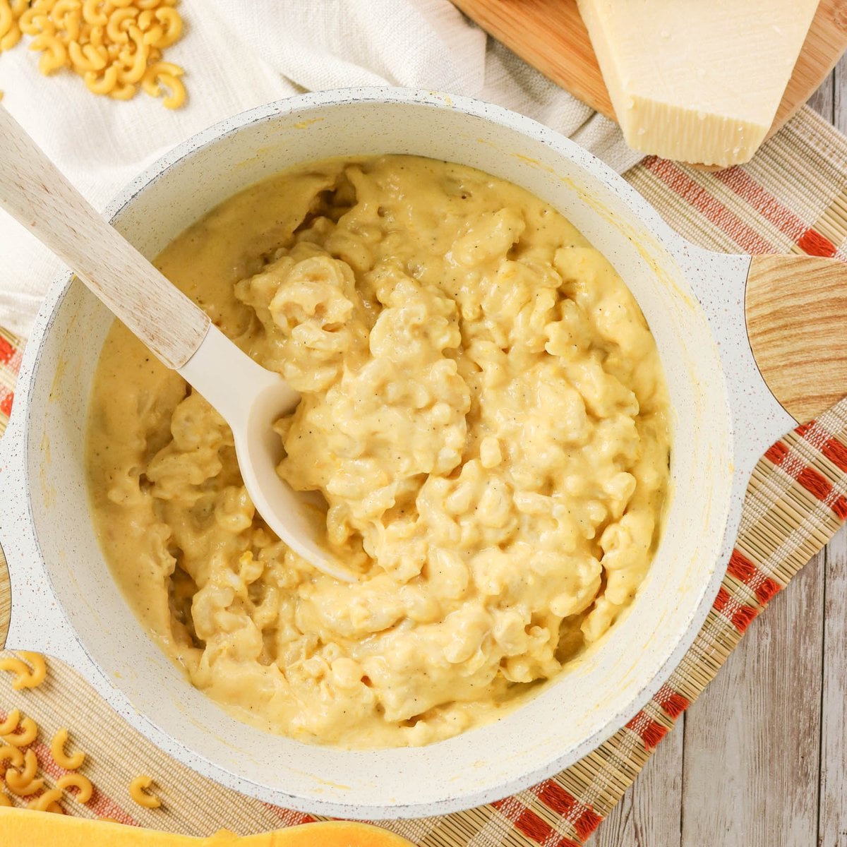 A pot of creamy macaroni and cheese with a white spoon, placed on a table with a block of cheese and uncooked macaroni nearby.