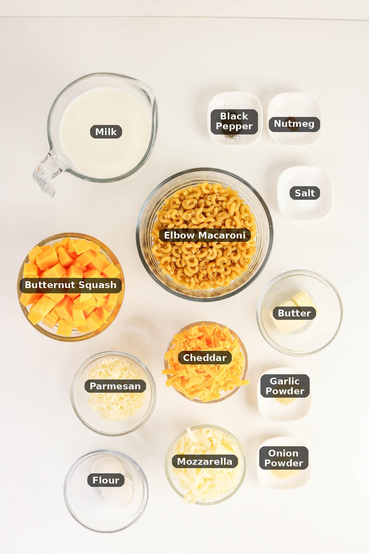 Top-down view of ingredients for macaroni and cheese, including elbow macaroni, butternut squash, cheese, milk, flour, butter, and seasonings in separate clear bowls.