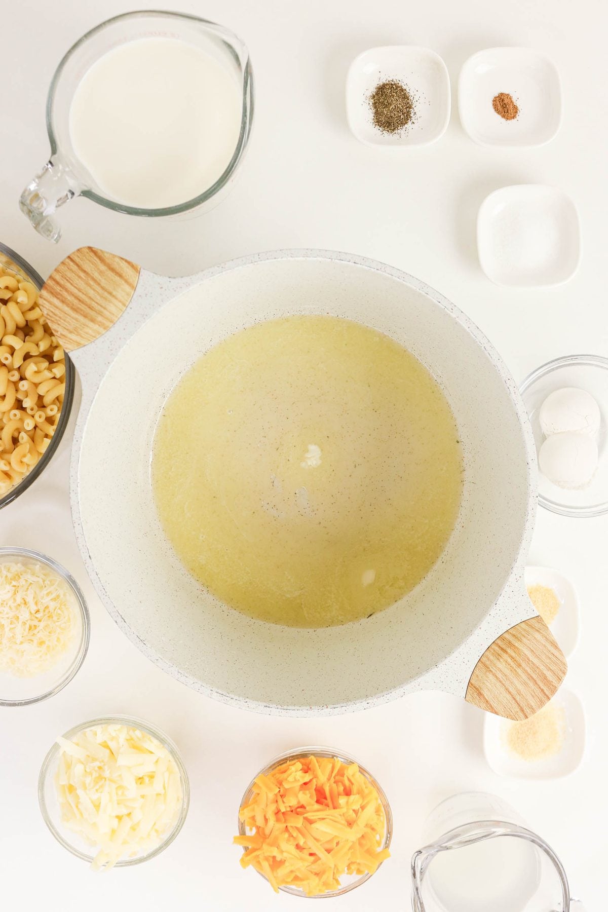 A pot of melted butter is surrounded by bowls of shredded cheese, uncooked pasta, milk, seasonings, eggs, and water on a white surface.