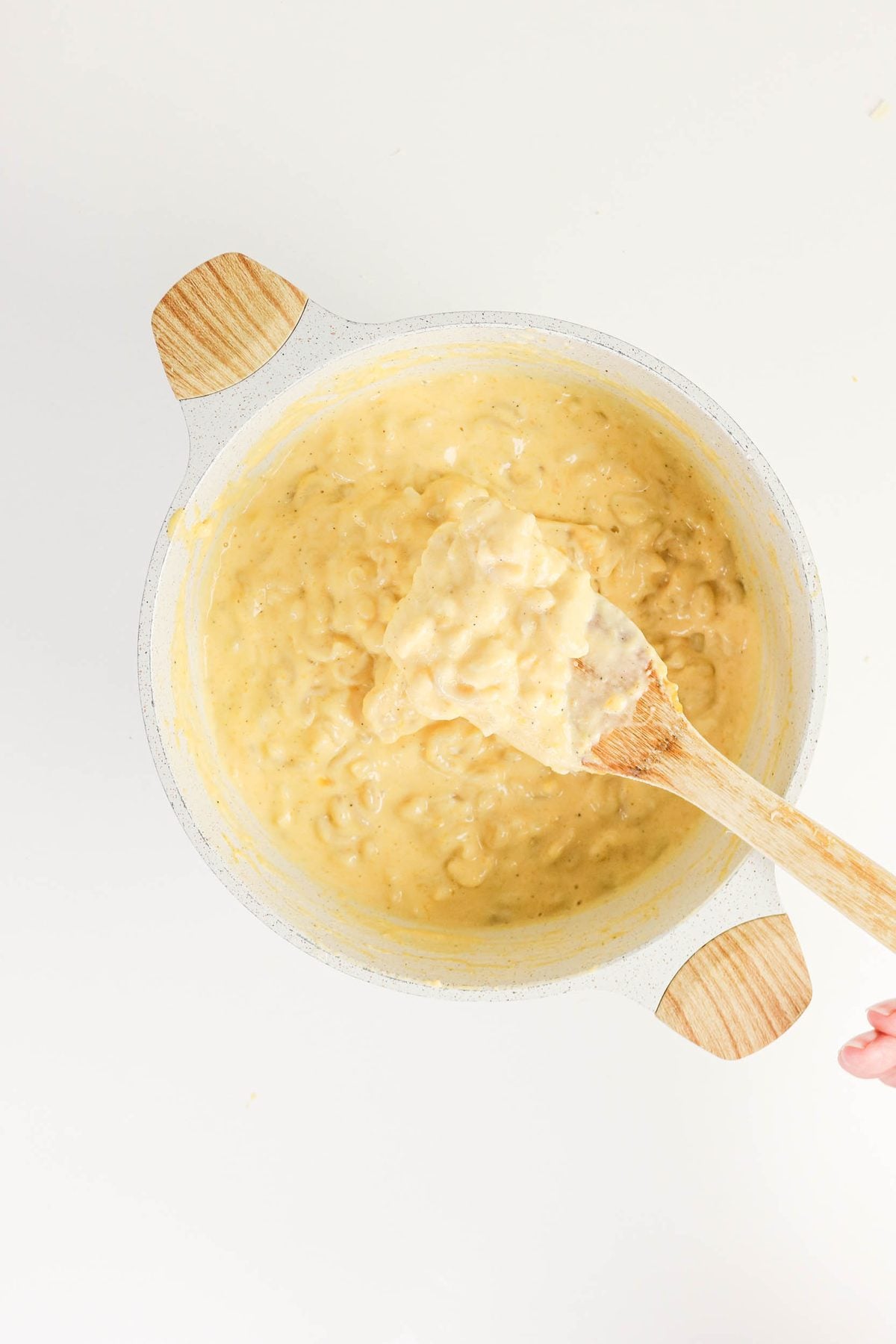 A pot of creamy macaroni and cheese is being stirred with a wooden spoon on a white surface.