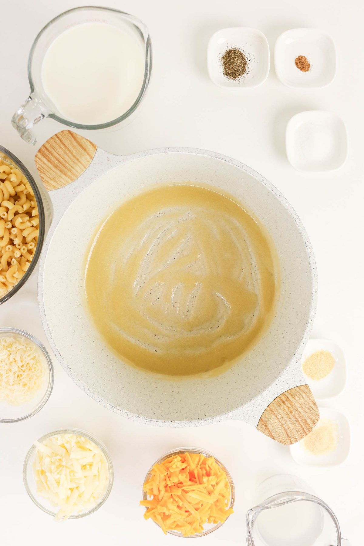 A white pot with melted butter is surrounded by bowls of shredded cheese, uncooked pasta, milk, and small dishes of spices on a white surface.