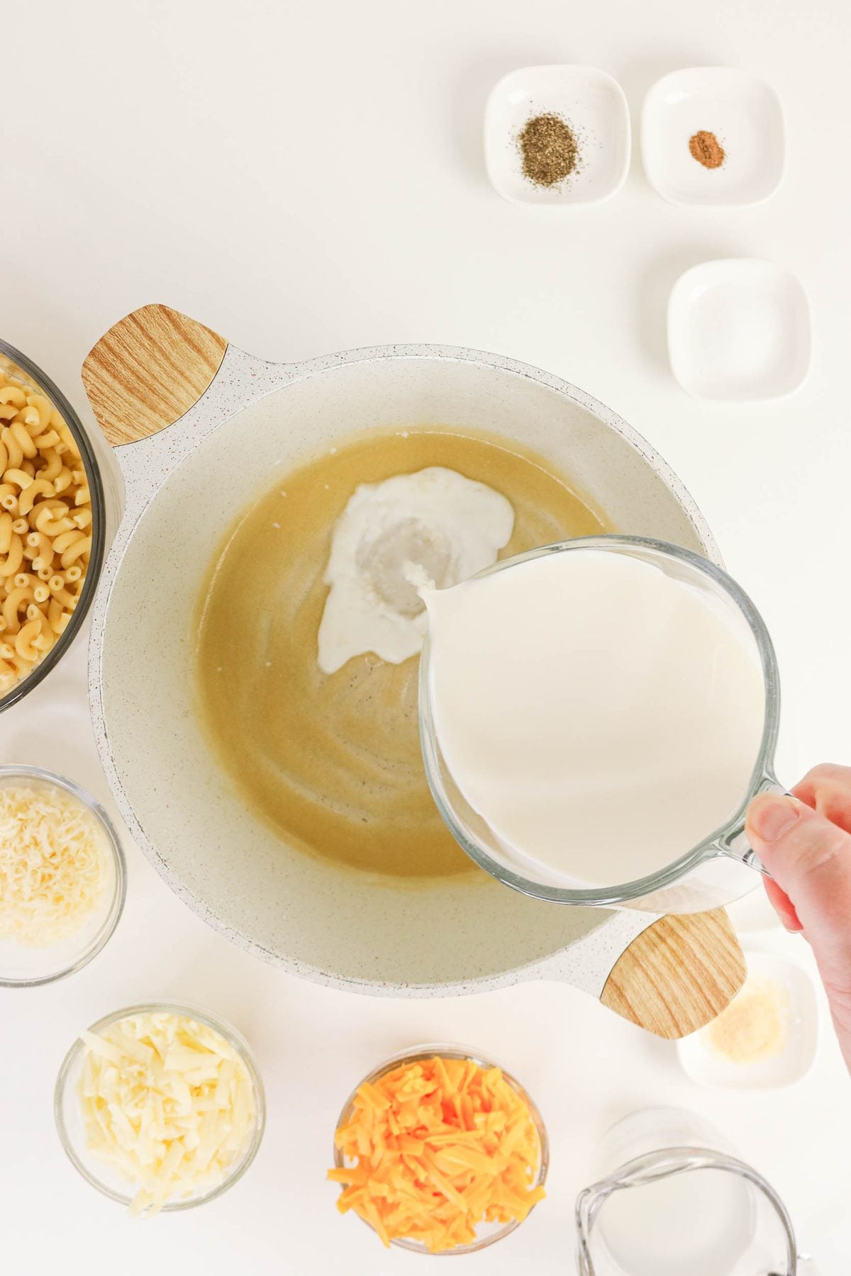 A hand pours milk from a glass measuring cup into a pot on a white surface, surrounded by bowls of pasta, shredded cheese, and small dishes of spices.