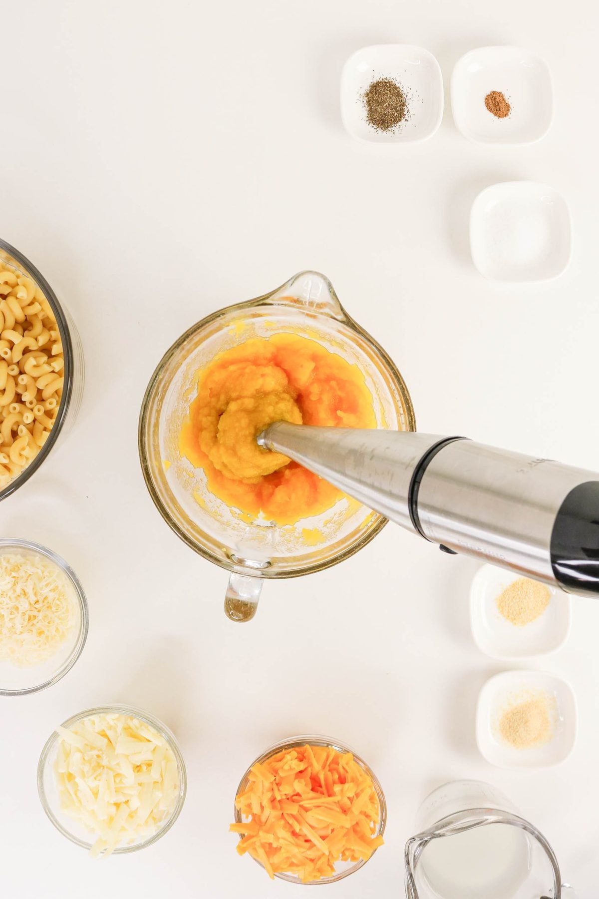 An immersion blender purees cooked squash in a glass bowl, surrounded by bowls of dry pasta, shredded cheese, spices, and a measuring cup of milk on a white surface.
