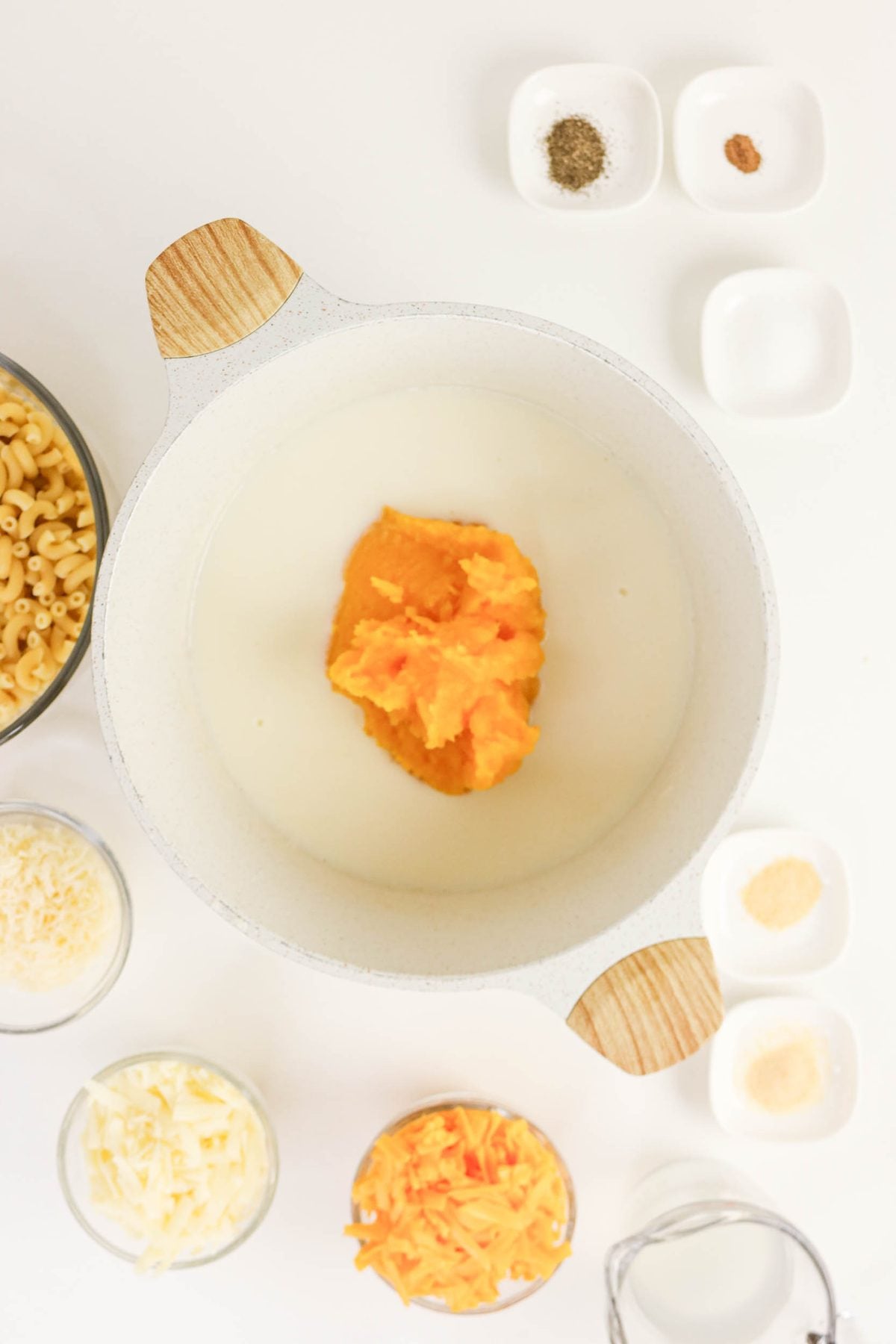 A white pot with milk and pumpkin puree, surrounded by bowls of shredded cheese, dry macaroni, spices, and milk on a white surface.
