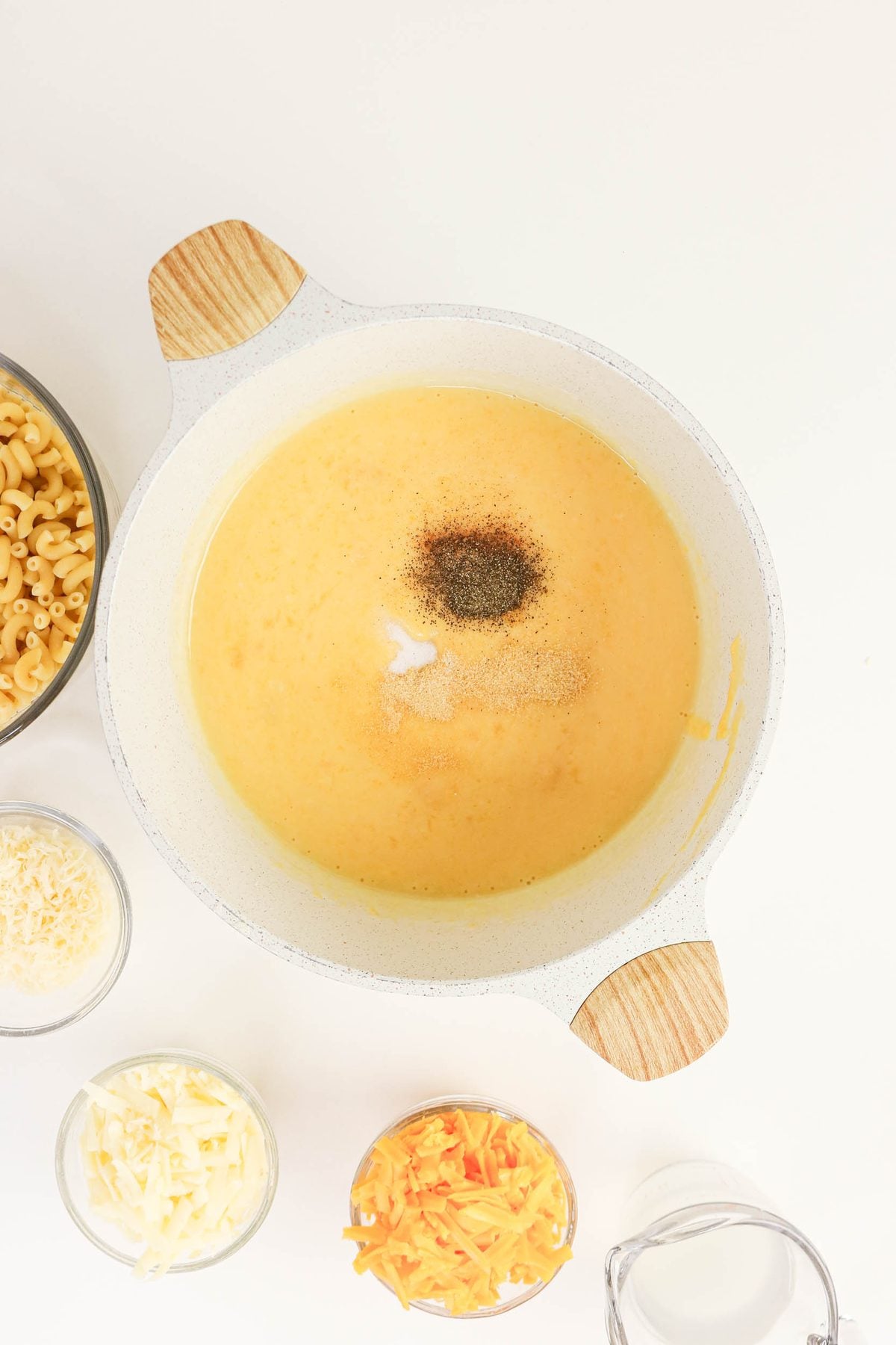 Large pot of cheese sauce with black pepper and salt on top, surrounded by bowls of uncooked pasta, shredded cheeses, and a cup of milk on a white surface.