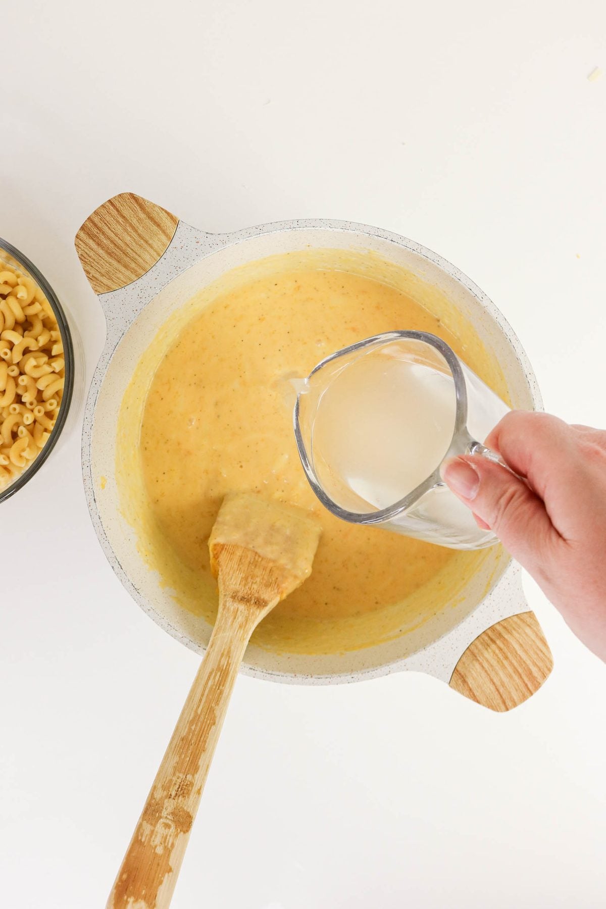A hand pours liquid from a measuring cup into a pot of cheese sauce with a wooden spatula inside. Cooked macaroni is visible in a bowl nearby.