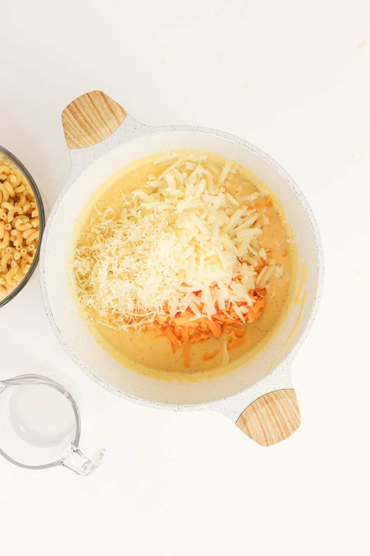 A pot with shredded cheese on top of a creamy sauce mixture, next to a bowl of uncooked macaroni and a glass measuring cup of milk on a white surface.