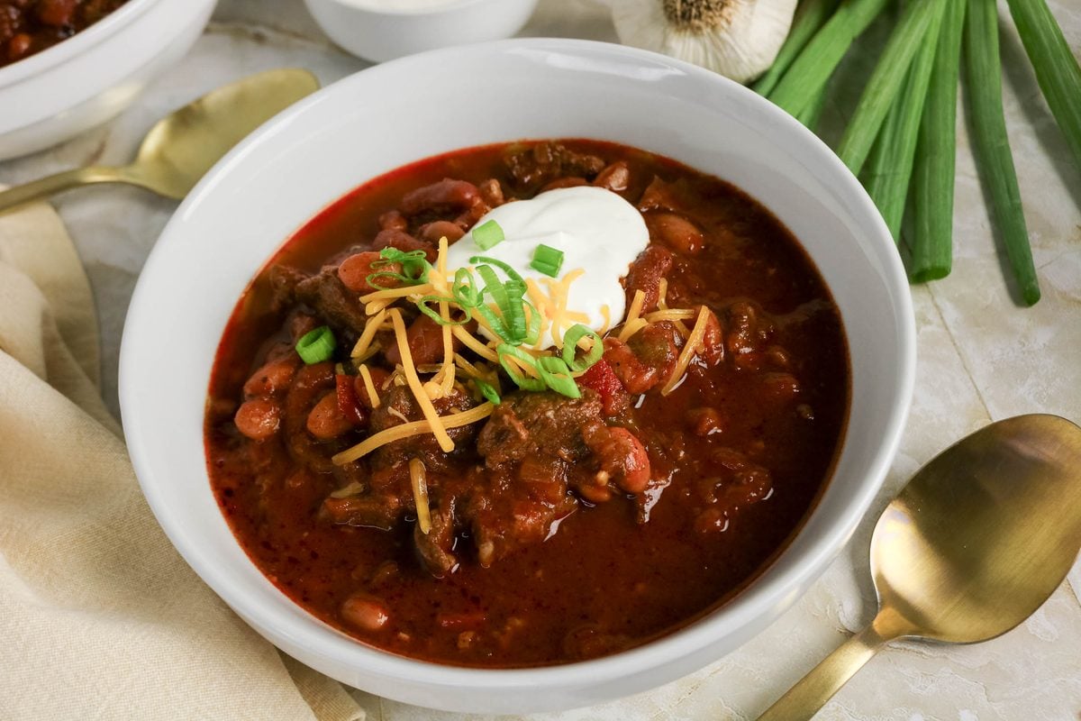 A bowl of chili topped with sour cream, shredded cheese, and sliced green onions, with a gold spoon and green onions nearby.