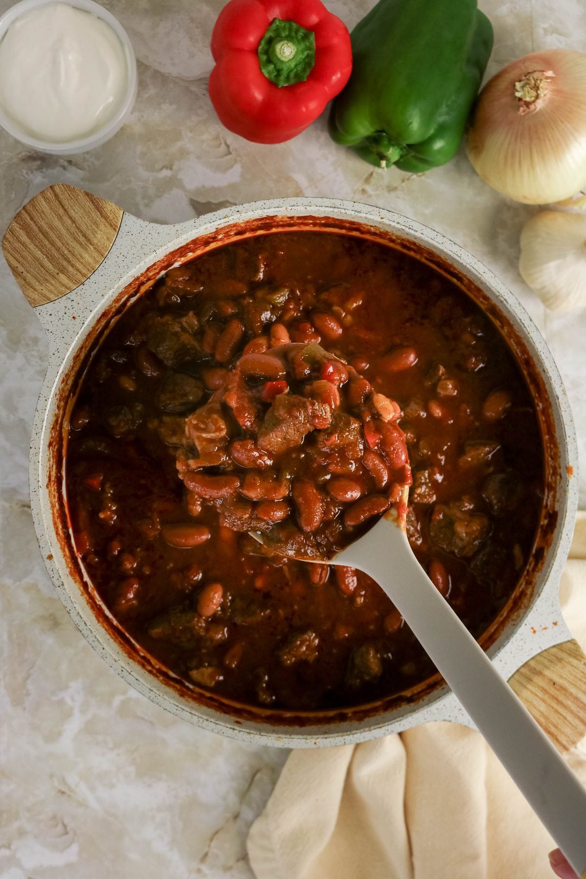 A pot of chili with beans and meat, with a ladle inside. Surrounding the pot are a red bell pepper, green bell pepper, onion, garlic, a bowl of sour cream, and a beige napkin.