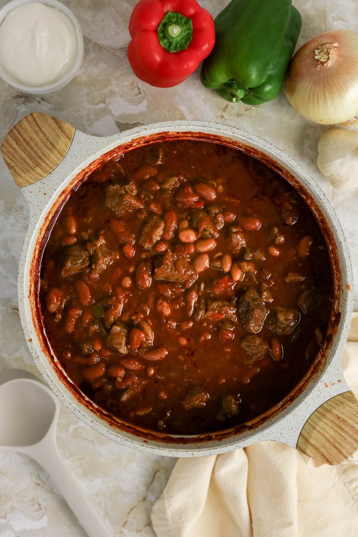 A pot of beef and bean chili sits on a countertop, surrounded by a red bell pepper, green bell pepper, onion, a bowl of sour cream, a ladle, and a kitchen towel.