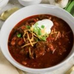 A bowl of chili topped with shredded cheese, sour cream, and chopped green onions, placed on a table with a garlic bulb and a spoon nearby.