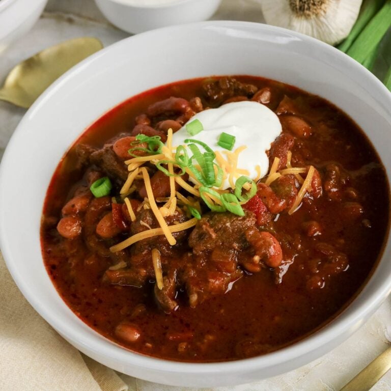 A bowl of chili topped with shredded cheese, sour cream, and chopped green onions, placed on a table with a garlic bulb and a spoon nearby.