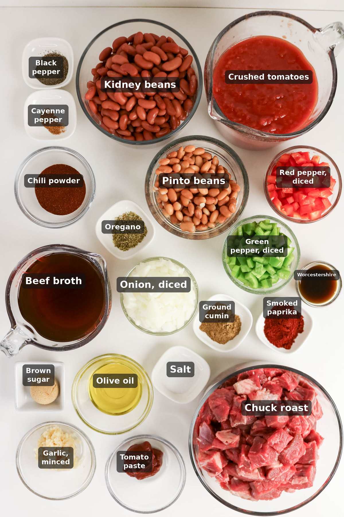 Ingredients for chili are arranged on a white surface, including beans, diced vegetables, spices, broth, olive oil, tomato paste, crushed tomatoes, garlic, and raw chuck roast.