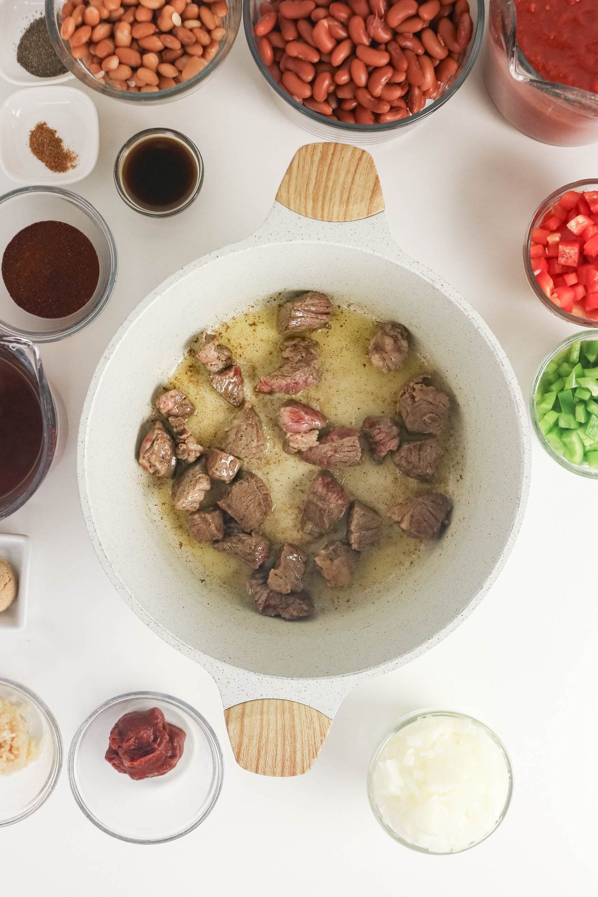Cubed beef browns in a white pot, surrounded by bowls of beans, chopped vegetables, spices, tomato paste, and sauce, arranged on a white surface.
